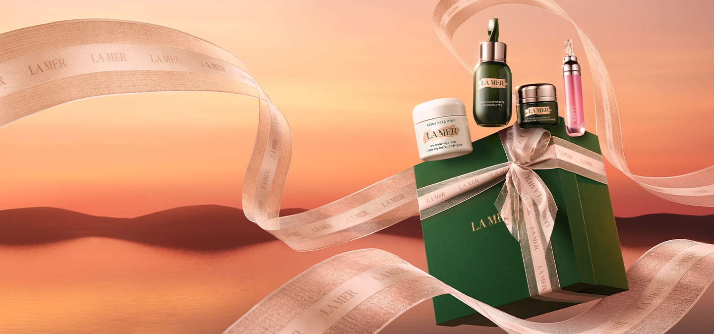 Green La Mer gift box with gold ribbon tied in a bow, with additional ribbon floating around and la mer products sitting on top.